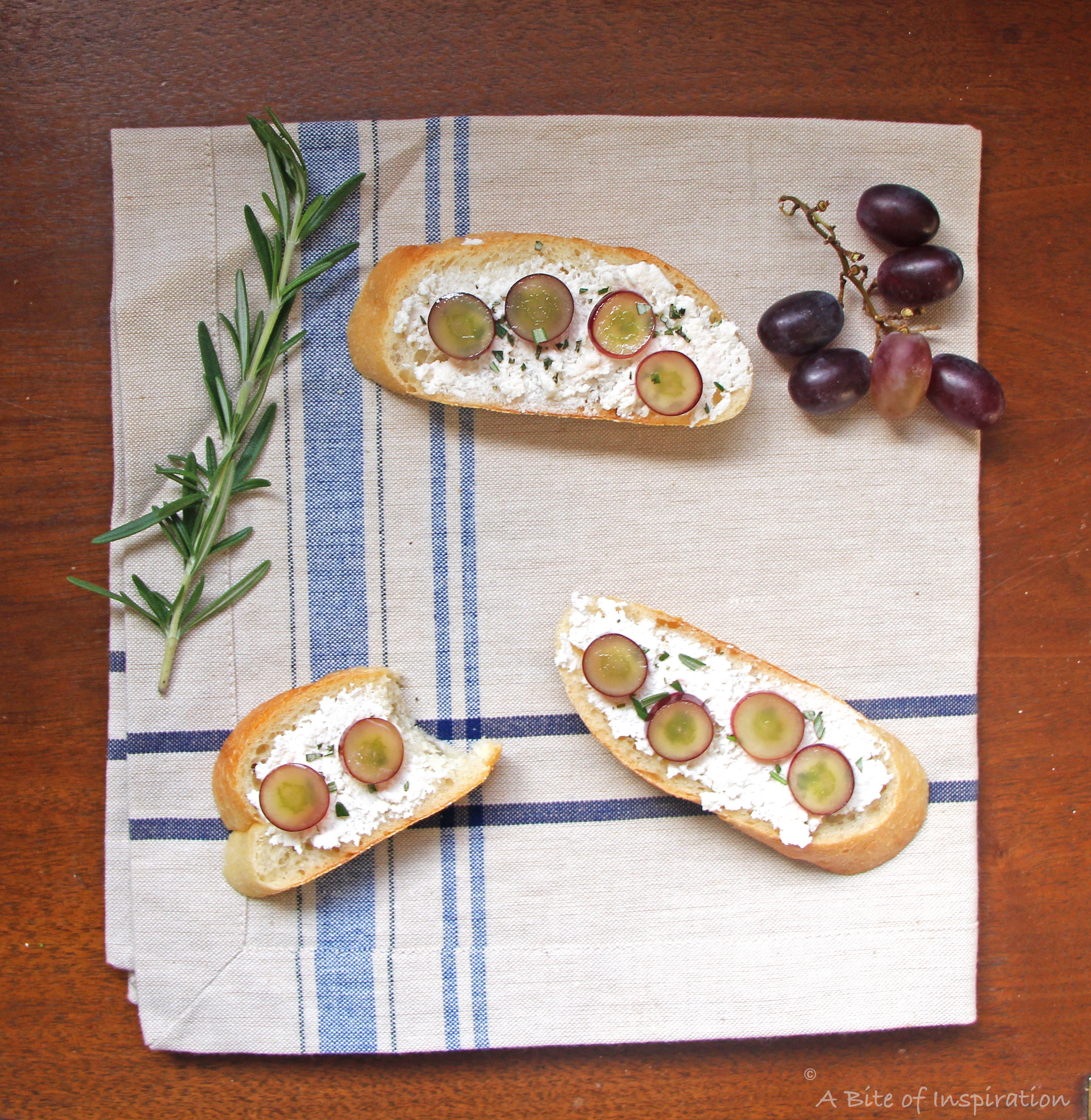 Three Soft Cheese Crostini with Pickled Grapes and Rosemary on a blue and white napkin with rosemary and grapes on the side