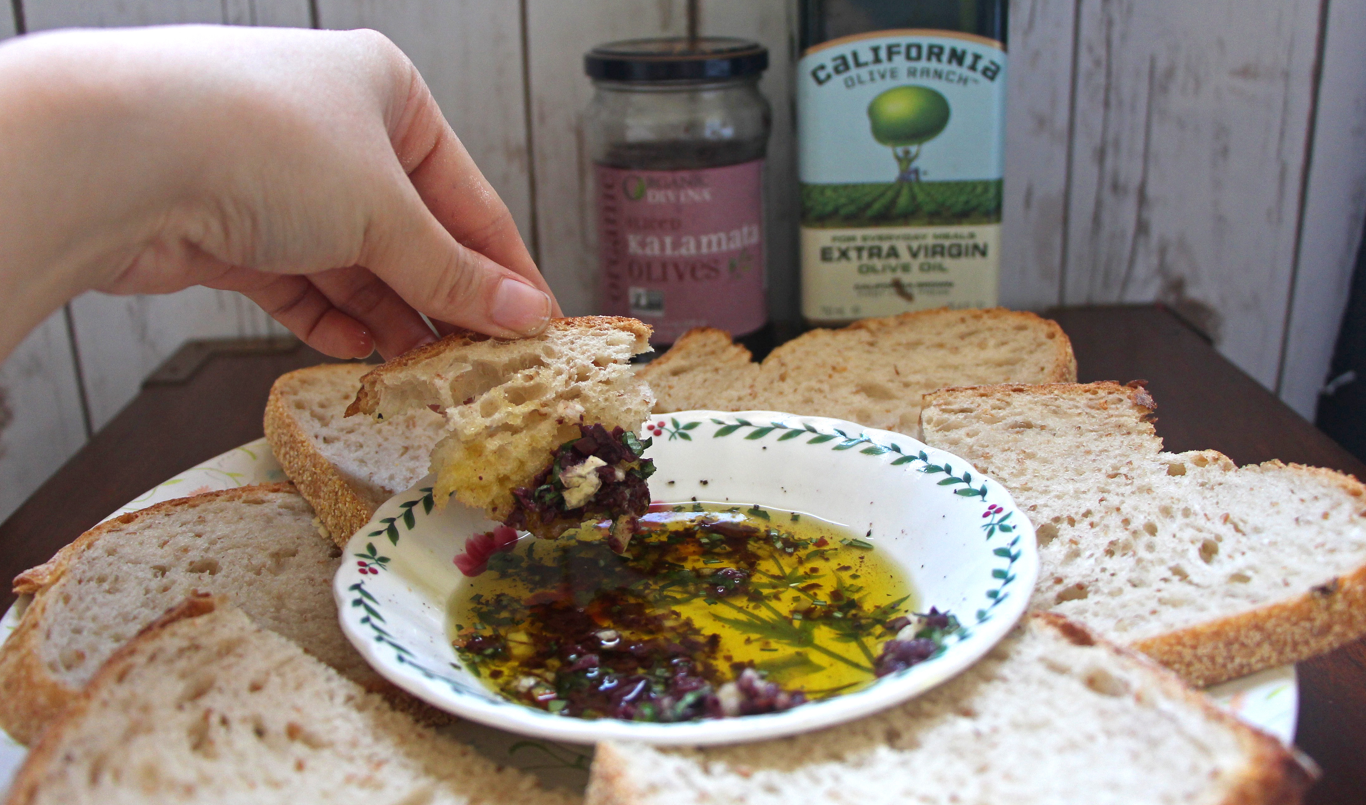 A piece of bread dipped into Olive Oil and Fresh Herb Sauce with kalamata olive jar and olive oil bottle in the background